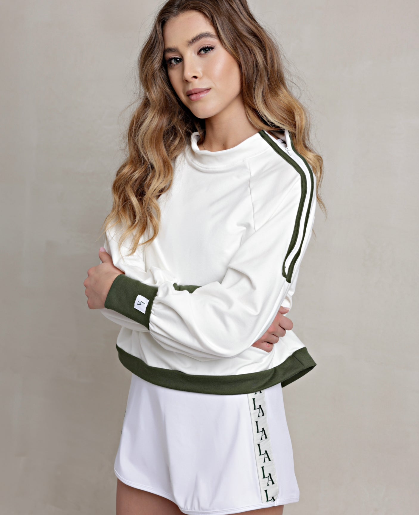 A female model wearing a white crewneck pullover with green trim on the waistband, cuffs, and shoulders. The pullover has a mock neck and zipper details on the sides.