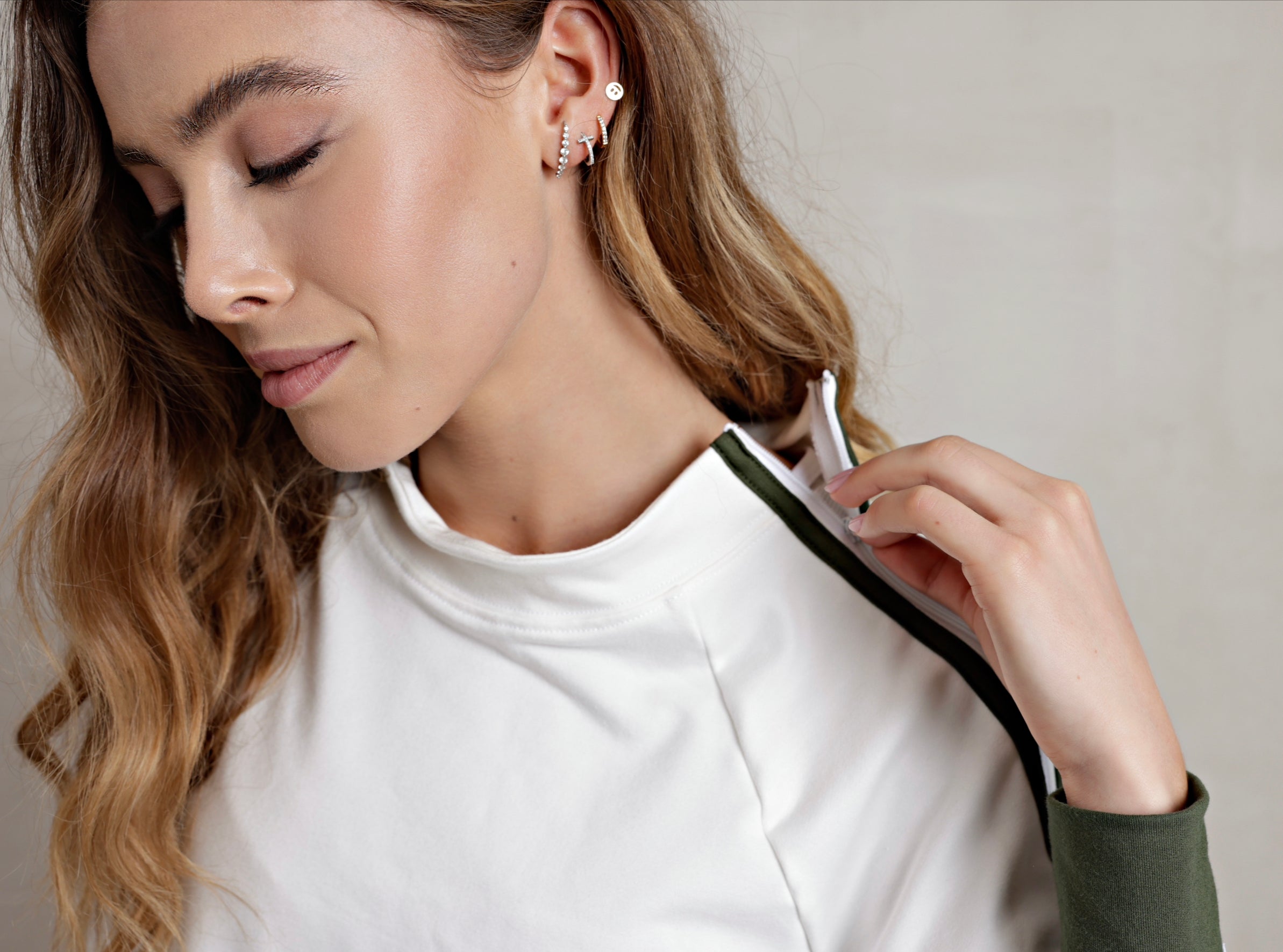 A female model wearing a white crewneck pullover with green trim on the waistband, cuffs, and shoulders. The pullover has a mock neck and zipper details on the sides. up close view