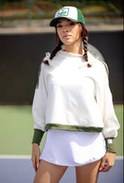 A female model wearing a white crewneck pullover with green trim on the waistband, cuffs, and shoulders. The pullover has a mock neck and zipper details on the sides.