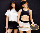 Two women wearing tennis attire, one holding a tennis racket and a ball, posing against a black background