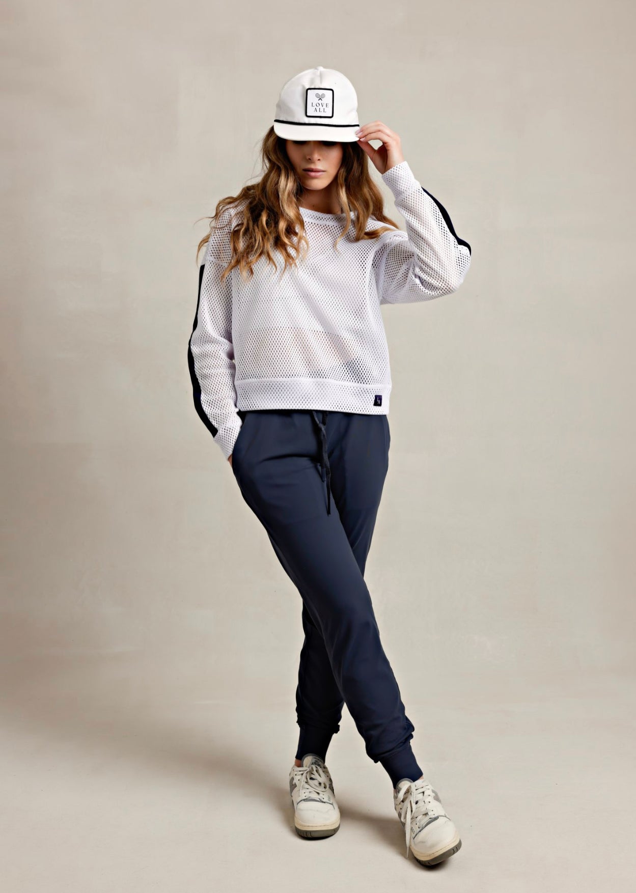 A woman wearing a white mesh pullover with navy snaps, paired with navy sweatpants, white sneakers, and a white cap.